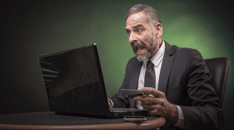 Mature man with a beard looking shocked while holding his credit card and looking at his computer